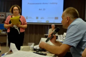 Mężczyzna siedzi przy stole i trzyma w dłoni mikrofon. W tle kobieta stoi z teczką. Za nią prezentacja multimedialna