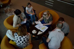 ludzie siedzą w fotelach w kole. Pochylają się nad stolikiem z broszurami. W tle kobieta na wózku inwalidzkim.