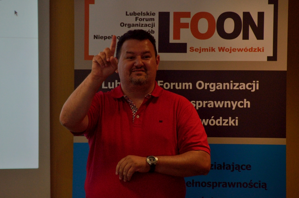 Mężczyzna stoi. Wyciąga dłon i palec wskazujący ku górze.