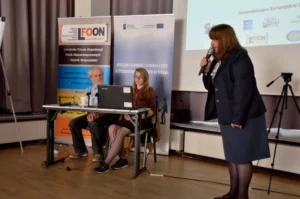 Kobieta trzyma mikrofon. W tle prezentacje multimedialne