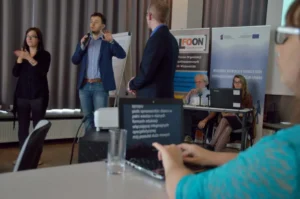 Ludzie stoja na środku. Mężczyzna ma mikrofon w dłoni.