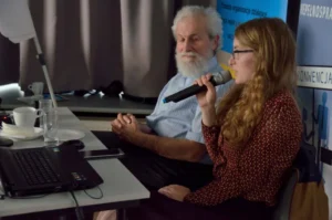 Kobieta siedzi. Trzyma mikrofon w dłoni.