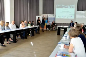 Kobieta trzyma mikrofon. W tle prezentacje multimedialne. Wokoło ludzie siedzący przy stołach.