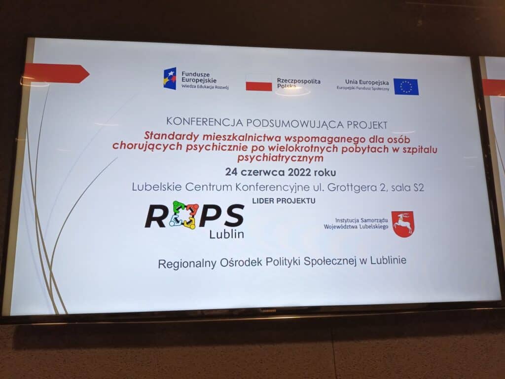 Zdjęcie slajdu tytułowego o treści: KONFERENCJA PODSUMOWUJĄCA PROJEKT: Standardy mieszkalnictwa wspomaganego dla osób chorujących psychicznie po wielokrotnych pobytach w szpitalu psychiatrycznym. 24 czerwca 2022 roku. Lubelskie Centrum Konferencyjne ul. Grottgera 2, sala S2. Lider Projektu: Pod tekstem: Logo ROPS Lublin, Logotyp Instytucji Samorządu Województwa Lubelskiego. Pod logami napis: Regionalny Ośrodek Polityki Społecznej w Lublinie