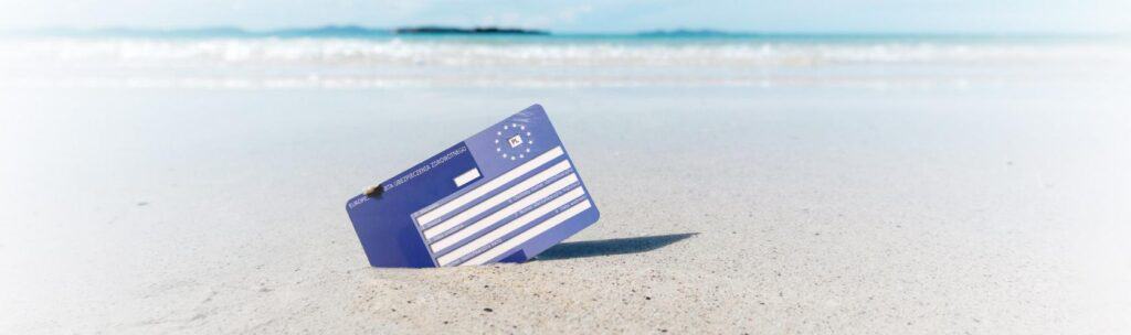 Karta EKUZ wbita w piasek na plaży. W tle błękitne morze.