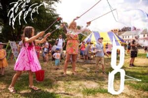 Kobieta i dziewczynka robią wielkie bańki mydlane. Obok kobiety stoi chłopczyk i przygląda się wielkiej bańce. W tle ludzie i dzieci patrzą na bańkowe arcydzieła.