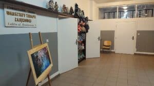 Korytarz z 3 drzwiami. Przed pierwszymi drzwiami na ścianie szyld: Stowarzyszenie "Źródło" Warsztaty Terapii Zajęciowej Lublin ul. Radzyńska 3. Pod nią sztaluga z ustawionym na niej obrazem. Przed drugimi drzwiami i obok pierwszych drzwi regał z wyrobami rękodzielniczymi.