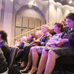 Publiczność siedząca na widowni, w usytuowanych „schodkowo” rzędach siedzeń; dużo ludzi różnej płci i w różnym wieku.
