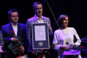 Od lewej stoi dwóch mężczyzn i jedna kobieta; mężczyzna w środku jest przedstawicielem firmy jubilerskiej „Savicki 1976 Sp. z o. o., ”uhonorowanej Medalem Prezydenta Miasta Lublin „W uznaniu za wieloletnią, pełną zaangażowania działalność na rzecz osób z niepełnosprawnościami oraz tworzenie miejsca służącego integracji i aktywizacji zawodowo-społecznej osób głuchych i słabo słyszących z podziękowaniem za szacunek i pomoc okazywaną drugiemu człowiekowi”.