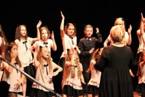 10 osób, stojąca tyłem krótkowłosa blondynka, ubrana w czarną tunikę - dyrygent chóru „Mała Kasjopea”, Pani Alicja Grzeszczyk, oraz dziewięcioro dzieci (8 dziewczynek ubranych w białe bluzeczki z czarnymi kokardkami i jeden ubrany na czarno chłopiec) podczas występu śpiewno-tanecznego