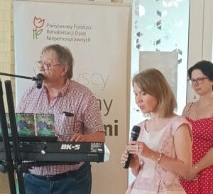 Mężczyzna z wąsem i w okularach stoi przy pianinie i przemawia do mikrofonu. Obok niego stoi blondynka z mikrofonem. Za blondynką brunetka w krótkich włosach