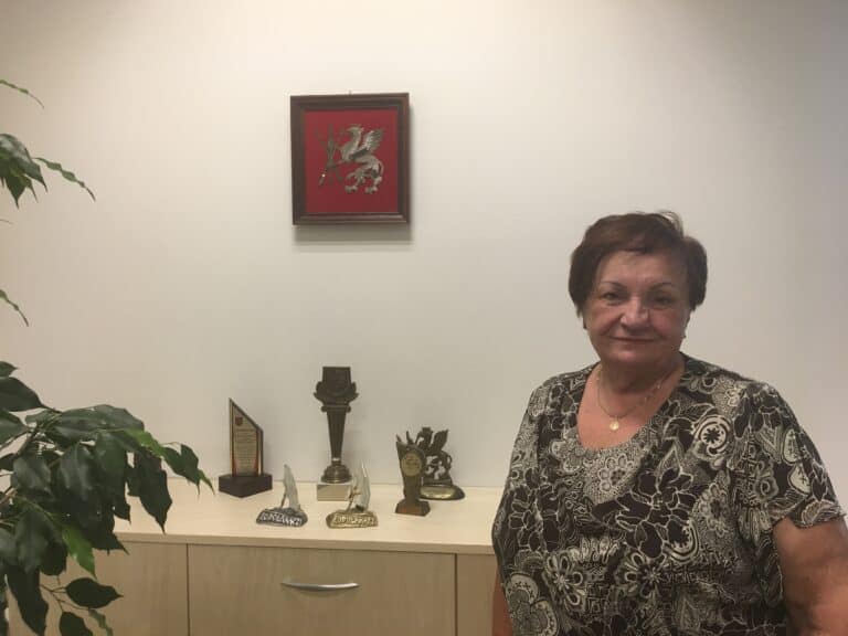 Stanisława Raczkiewicz, starsza kobieta z ciemnymi krótkimi włosami, ubrana we wzorzystą czarno-beżową sukienkę stoi obok komody, na której ustawione są przyznane statuetki.