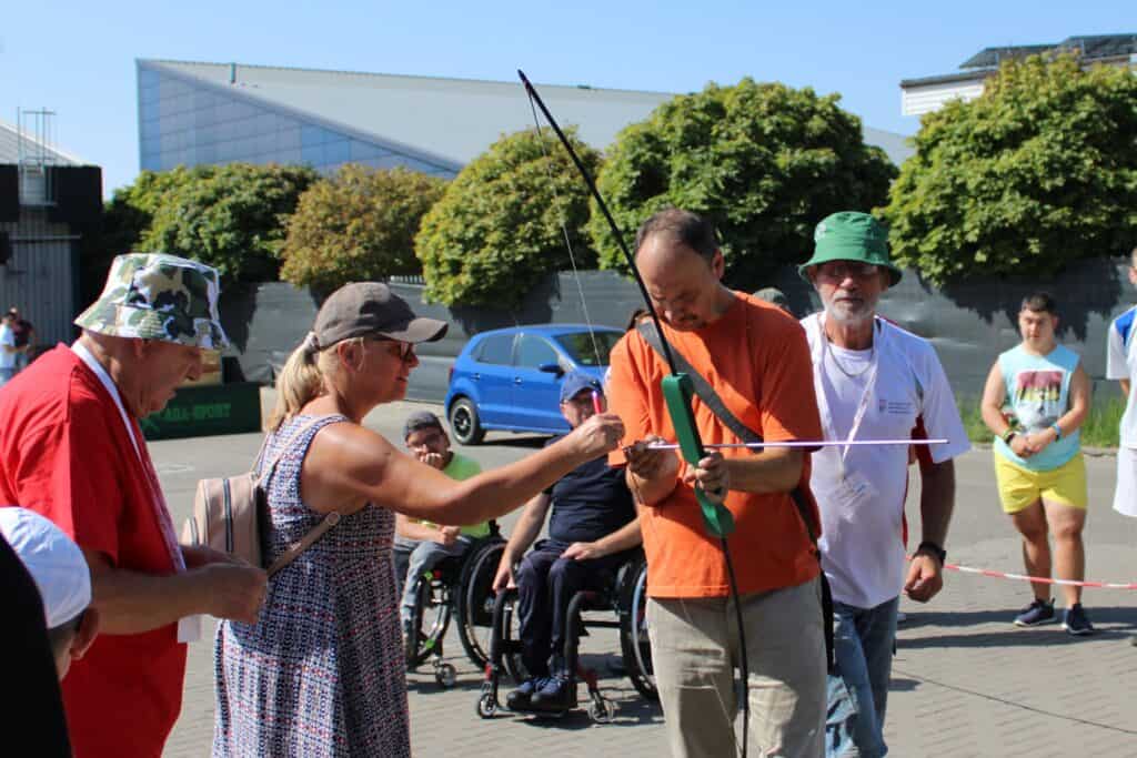 Zawodnik podczas przygotowania do konkurencji strzelanie z łuku w III Dniu Pikniku. W tle obserwujące osoby w tym dwie na wózku inwalidzkim