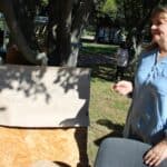 Kobieta w średnim wieku stoi obok płyty pilśniowej opartej o drzewo. Na płycie arkusz z napisem Psycholog p. Kasia