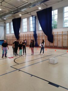Sportowcy wraz z kobietą sędzią trzymającą listę stoją na środku sali gimnastycznej. Jedna z uczestników przygotowuje się do rzutu woreczkiem do białej skrzynki ustawionej w pewnej odległości. W kolejności oczekują pozostali uczestnicy.