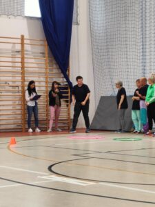 Dwie kobiety sędzie, jedna z listą, druga z gwizdkiem przy ustach i stoperem stoją przy drabince. W kierunku egzaminatorek wychyla się sportowiec. A za nim oczekujące cztery osoby. Na podłodze znajdują się dwa koła hula hop jedno duże i różowe, drugie małe i zielone.