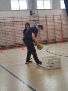 Sportowiec pochyla się z piłką w ręku. Obok stoi skrzynka pomalowana na biało. W tle przypatrująca się jemu kobieta.