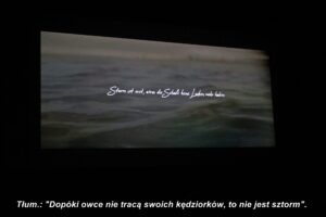 Zbliżenie na ekran kinowy, wyswietlający fale morskie. Na środku fali napis: Sturm ist erst wenn die Schafe keine Locken mehr haben. Do zdjęcia dodany dopisek: Tłum.: "Dopóki owce nie tracą swoich kędziorków, to nie jest sztorm".