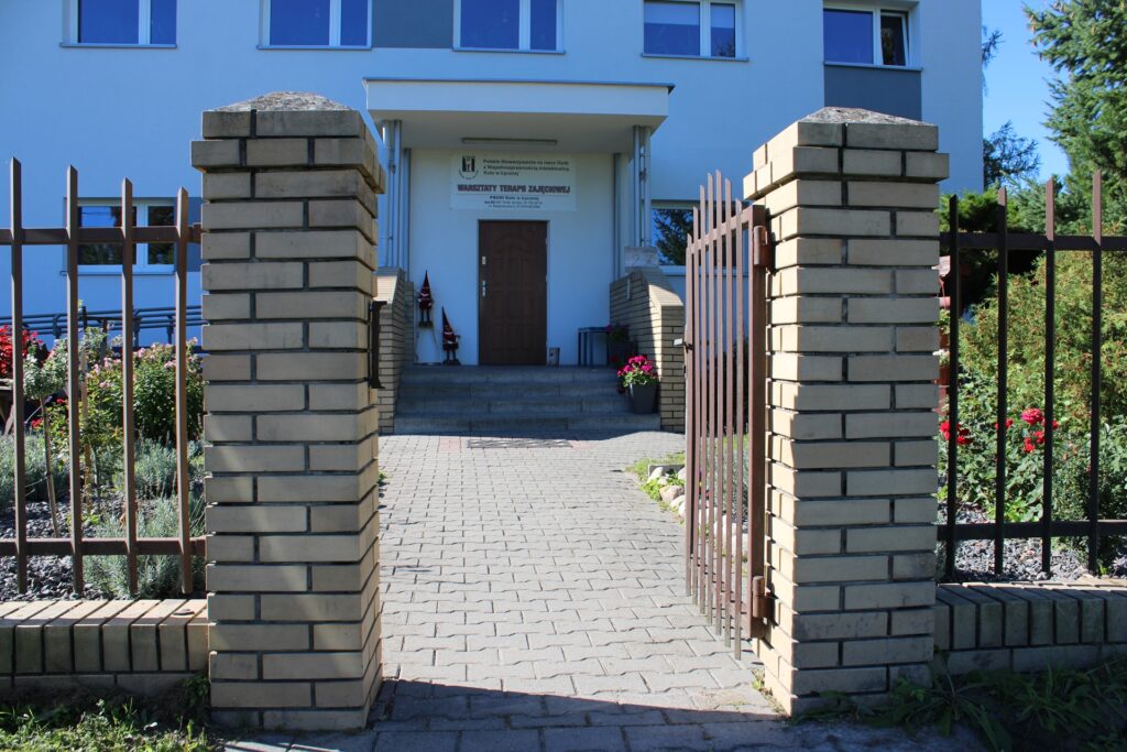 Furtka ogrodzeniowa z wejściem do budynku na którym widnieje szyld Warsztaty Terapii Zajęciowej PSONI Koło w Łęcznej.