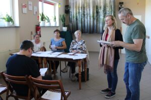 Trzy panie w średnim wieku siedzą przy stole na których leżą rozłożone dokumenty. Obok nich stoi młoda kobieta z mężczyzną w średnim wieku. Tyłem do aparatu i na wprost trzech pań siedzących siedzący mężczyzna w okularach.