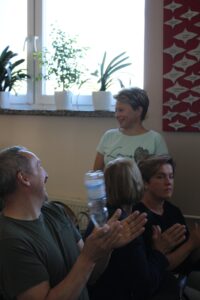 Siedzące dwie kobiety i mężczyzna zwróceni są w kierunku stojącej za nimi kobietę klaszczą w ręce. Mężczyzna trzyma w ręku odwróconą do góry dnem buteleczkę wody.