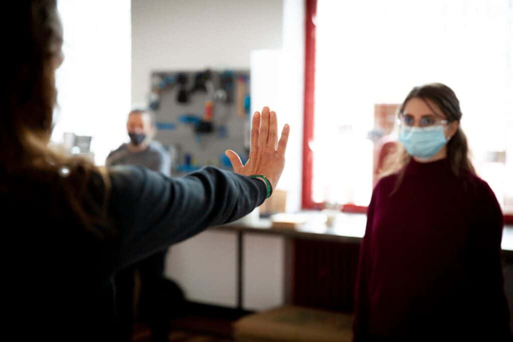 Kobieta zwrócona tyłem do aparatu pokazuje stojącej naprzeciwko niej kobiecie w maseczce wyprostowaną rękę z dłonią odchyloną ku górze