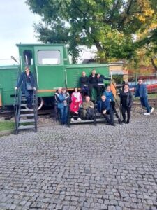 Grupa 13 osób pozuje do zdjęcia z zieloną lokomotywą w tle. Cztery osoby opierają się o ścianę boczną lokomotywy, część siedzi na schodkach, część stoi.
