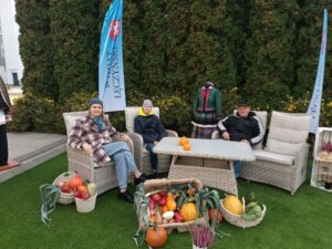 Dwie kobiety i jeden mężczyzna siedzą na fotelach ogrodowych z technorattanu. Na stole leżą dwie dynie. Przed nimi ustawione są koszyki z kolorowymi warzywami.