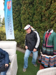 Mężczyzna w białym berecie pozuje obok manekina bez głowy prezentującego kobiecy strój ludowy. W tle żywopłot i flaga reklamowa z napisem Powiat Łęczyński.