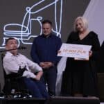 Siedzący na elektrycznym wózku inwalidzkim Pan Patryk Grab – stypendysta II edycji Funduszu Wieczystego im. Piotra Pawłowskiego podczas wręczenia przez Panią Ewę Pawłowską – Prezes Fundacji Integracja oraz Pana Pawła Łukasiaka – Prezesa Akademii Rozwoju Filantropii w Polsce, symbolicznego czeku na sumę 10 000 zł
