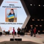 Cztery osoby stojące na scenie, od lewej Pani Katarzyna Cimoch z Fundacji Kulawa Warszawa, Pani Ewa Pawłowska – Prezes Fundacji Integracja, Pan Tomasz Wolny i kobieta w czerwonej sukience, za nimi na sześciokątnym ekranie portret Pani Izabeli Sopalskiej-Rybak – Prezes Fundacji Kulawa Warszawa, uhonorowanej Medalem „Przyjaciel Integracji”