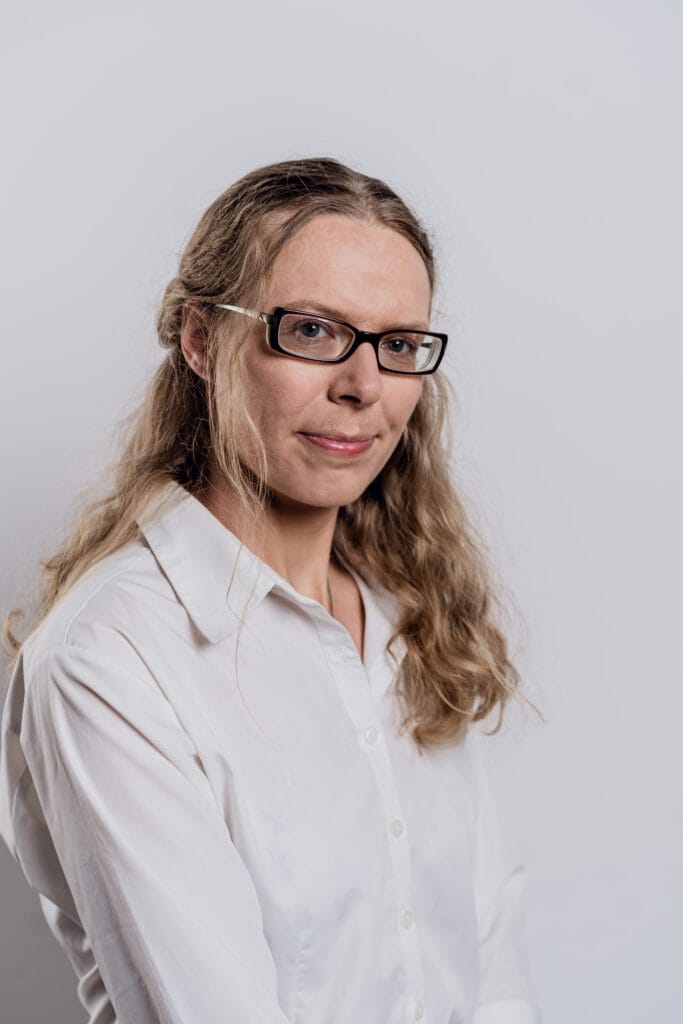 Alena Androsik, młoda kobieta długimi blond włosami, w okularach z uśiechem patrzy w obiektyw. Ubrana w białą rozpiętą pod szyją koszulę. Zdjęcie portretowe.