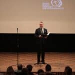 Stojący na scenie przy mikrofonie Pan Jakub Semeniuk - Kierownik Warsztatów Terapii Zajęciowej w Białej Podlaskiej, podczas powitania zgromadzonej w Sali kinowej publiczności