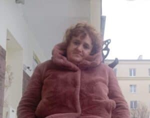 Renata Tkaczyk - kobieta w średnim wieku, kręcone włosy, ubrana w futerko w kolorze pudrowy róż. Z delikatnym uśmiechem patrzy w obiektyw