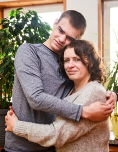 Anna Chwałek - kobieta w średnim wieku z długimi kręconymi włosami, uśmiechnięta - stoi przytulona do swojego syna Kuby - młodego mężczyzny, wyższego od mamy. ich głowy są do siebie przytulone. Mam z synem obejmują się nawzajem.