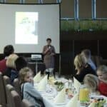 Na pierwszym planie siedzące przy zastawionych stołach rodziny z dziećmi uczestniczące w Projekcie „Rodzinne Wędrówki”; w tle od lewej stoją: Elżbieta Wiśniewska (Koordynator Projektu „Rodzinne Wędrówki”), Elżbieta Krupa (Wiceprezes Stowarzyszenia Synergia), Aleksandra Bednarz (Psycholog)