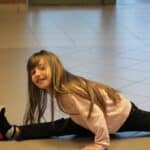 Dziewczynka wykonująca tzw. szpagat francuski, tj. figurę gimnastyczną, w której jedna noga jest wystawiona w przód, a druga w tył, tworząc linię prostą wzdłuż podłogi