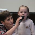 Elżbieta Krupa (Wiceprezes Stowarzyszenia Synergia) przytrzymująca mikrofon śpiewającej do niego dziewczynce