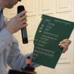 Zbliżenie na trzymany w ręku uszkodzony fragment ciemnozielonej tablicy informacyjnej „Budynek CEUE poziom 3”, zawierającej kolorowe piktogramy, białe napisy oraz odpowiadające im oznaczenia w alfabecie Braille'a