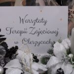 Biała kartka wielkości A4 z czarnym napisem „Warsztaty Terapii Zajęciowej w Oleszycach”, umieszczona pomiędzy świątecznymi dekoracjami rękodzielniczymi, przeznaczonymi do sprzedaży podczas Mikołajkowego Jarmarku Pracy