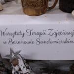 Biała kartka wielkości A4 z czarnym napisem „Warsztat Terapii Zajęciowej w Baranowie Sandomierskim”, umieszczona pomiędzy świątecznymi dekoracjami rękodzielniczymi, przeznaczonymi do sprzedaży podczas Mikołajkowego Jarmarku Pracy