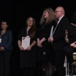 Na scenie stoją Przedstawiciele, odbierających podziękowania Warsztatów Terapii Zajęciowej: trzy kobiety i ks. Tomasz Szostek, po prawej stronie na wysokim krześle siedzi Katarzyna Hul (Tłumacz PJM)