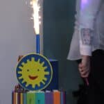 Dwupiętrowy tort z odpaloną świetlną racą, przyozdobiony symbolem uśmiechniętego słońca, oraz wykonanymi z czekolady kredkami i książkami