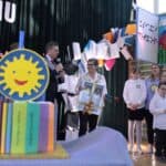 Na pierwszym planie dwupiętrowy tort, przyozdobiony symbolem uśmiechniętego słońca, oraz wykonanymi z czekolady kredkami i książkami a w tle Bożena Kazanowska - laureatka Orderu Uśmiechu w towarzystwie Kanclerza, Członków Kapituły oraz uczniów SOSW