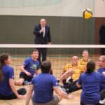 Dziewięciu zawodników obu płci z obu drużyn patrzy w górę na lecącą piłkę w oddali stoi komentator i dwóch kibiców