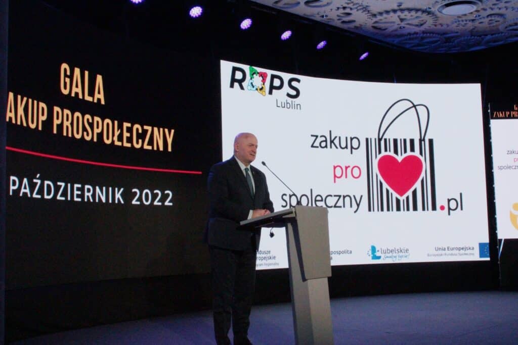 Pan Jarosław Stawiarski, Marszałek Województwa Lubelskiego, łysy mężczyzna w średnim wieku ubrany w ciemny garnitur, białą koszulę i krawat, stojący przy mównicy w tle za jego plecami czarny ekran z żółtym napisem „Gala Zakup Prospołeczny” pod czerwoną poziomą kreską biały napis „14 październik 2022”, po prawej stronie na sąsiednim ekranie biała plansza z logo zakup pro społeczny, składającym się z czarnego kodu kreskowego z rączkami, przypominającego torbę na zakupy, w środku, której umieszczono czerwone serce.