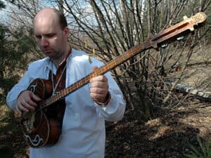 Bohater wywiadu ubrany w ludową koszulę z haftem przy dekolcie z instrumentem -tuszpulurem, wschodnim trzystrunowym instrumentem z republiki Tuwy. Wyglądający jak mała gitara z długim gryfem lub płaskie jabłko z długim ogonkiem