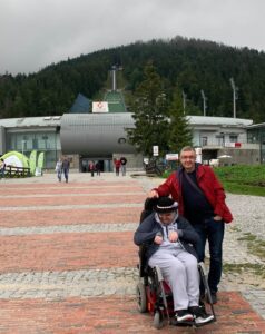 Mateusz Bobruś - młody mężczyzna na wózku ubrany w szary dres i grafitowy dres oraz góralski kapelusz z piórkiem. Obok stoi Jacek Bobruś- wysoki mężczyzna w czerwonej kurtce i granatowych dżinsach. Panowie stoją na tle skoczni narciarskiej w Zakopanem.