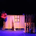 Scenografia w spektaklu pt. „Zupa z kamienia”: parawan z okienkiem i fragment drewnianego ogrodzenia, stylizowane na wiejską chałupę