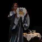 Scena w spektaklu pt. „Chory Kotek”: kobieta ubrana w długą, powłóczystą, srebrną szatę, trzyma w rękach rysunek myszy - symbolizuje marzenia chorego kotka o smakołykach